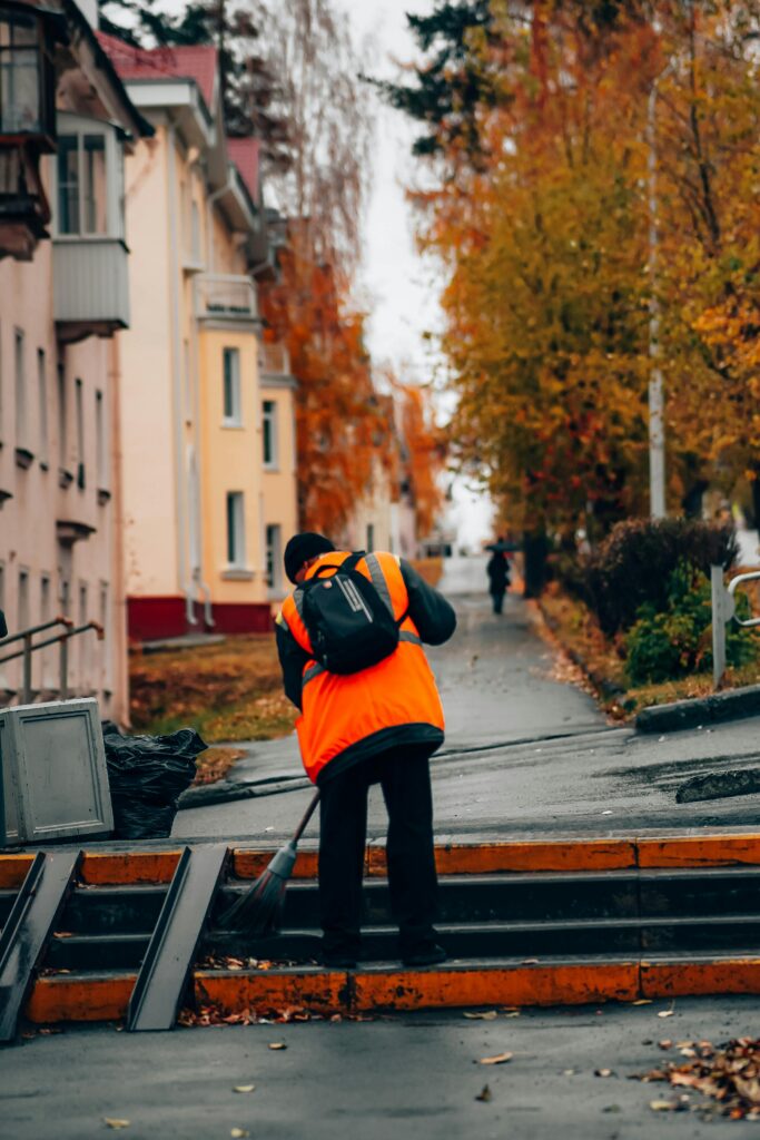 A man cleaning the sidewalk with a broom during autumn in a city neighborhood.
