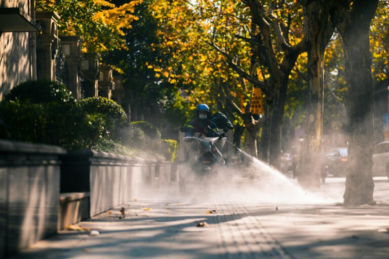 A city worker sprays a sidewalk with water on a sunny day, surrounded by lush trees and buildings.