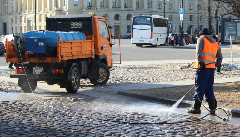 A city worker uses a water hose to clean the urban streets in a sunlit scene.